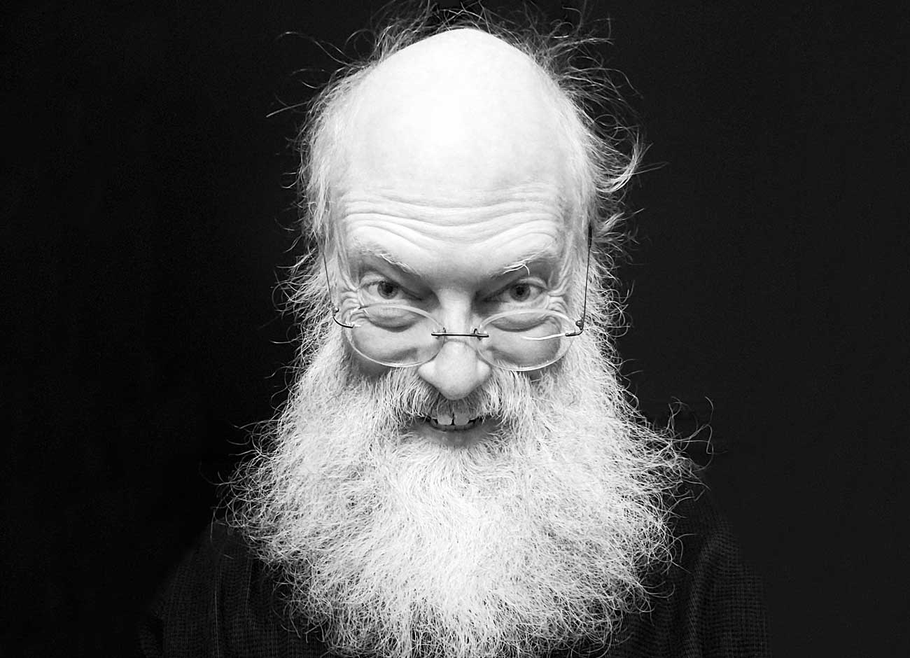 a black and white image of a 52 year old man smiling and wearing glasses. He has a long wispy white beard
