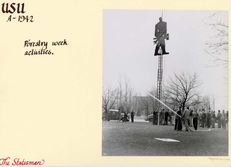 A black and white image from 1942 of a wooden Paul Bunyan statue atop a ladder. A group of men is using a firehose to keep people away while they figure out how to get Bunyan down.