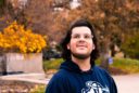 a smiling young man wears an Aggie sweatshirt and looks up at the sky with fall colors behind him