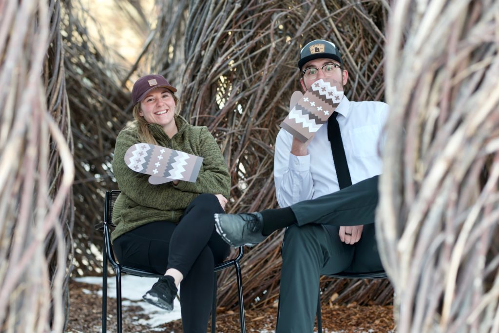 two students sit on chairs outside wearing oversized mittens like the Bernie Sanders internet meme and grinning