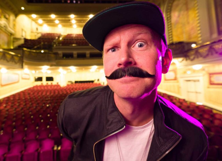 a man sits in an empty theater. he is wearing a fake mustache and amused expression