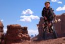 a young man in traditional Navajo dress stands on a sandstone rock and holding a bow and arrow with the backdrop of Monument Valley Utah