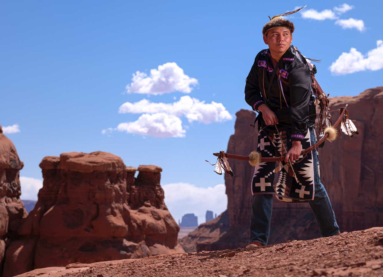 a young man in traditional Navajo dress stands on a sandstone rock and holding a bow and arrow with the backdrop of Monument Valley Utah