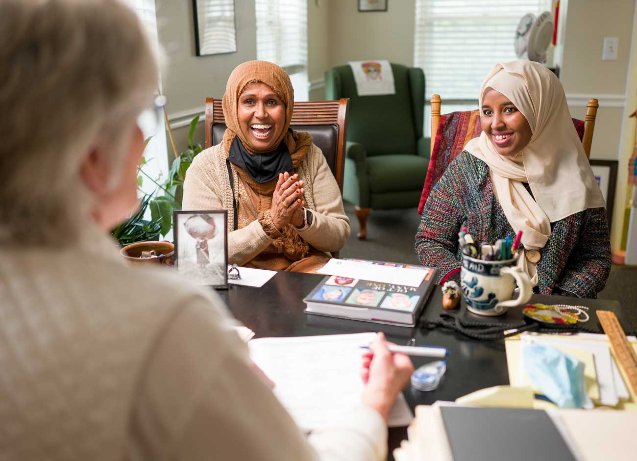 two women sit in an office smiling while talking to another woman across a table. The two women are of African descent and wearing head scarves. A pile of paperwork sits on the desk.