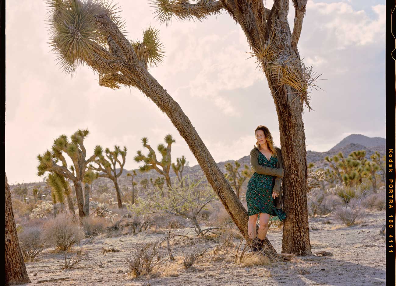 a woman in a windblown green dress and sweater leans against a Joshua tree in the desert. The sky is a pale pink color.
