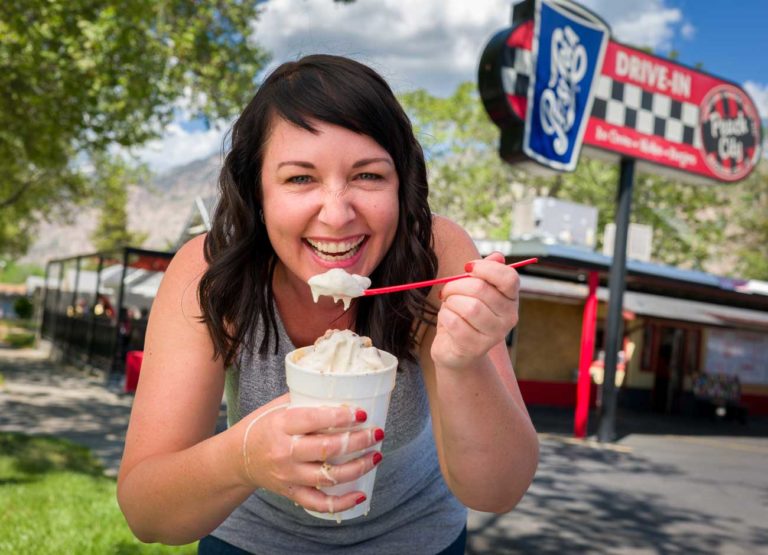a woman with black hair grins while holding a melting milkshake and eating it with a spoon. the drips fall down her hands. she stands in front of a drive in.