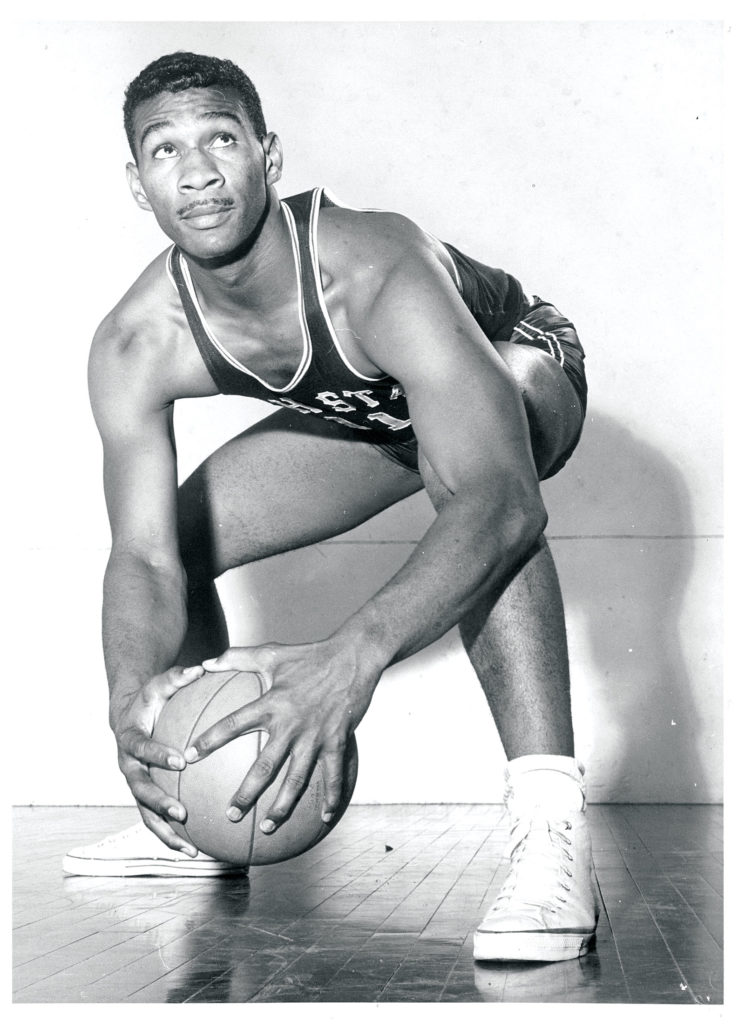 An African American basketball player squats holding a basketball. His eyes gaze upward as if sizing up his next shot.