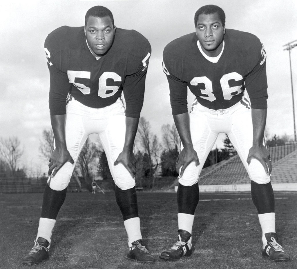 Two African American men in football uniforms stare into the distance. Both have their hands on their knees.
