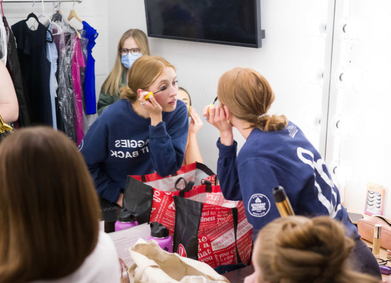 a young woman leans toward a large mirror as she puts on her makeup. a rack of evening dresses is in the background. she is wearing an Aggie t-shirt.