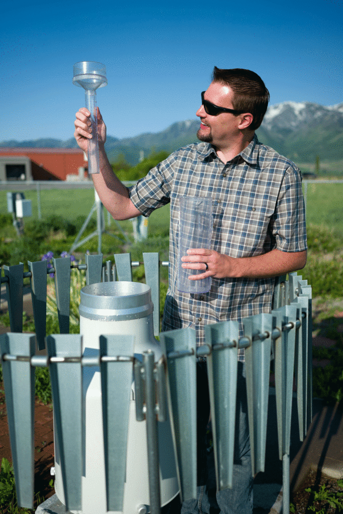 a man stands in the  center of a high-tech metal circular rain gauge. He is wearing sunglasses and holding a small low-cost version of the rain gauge used by citizen scientists.