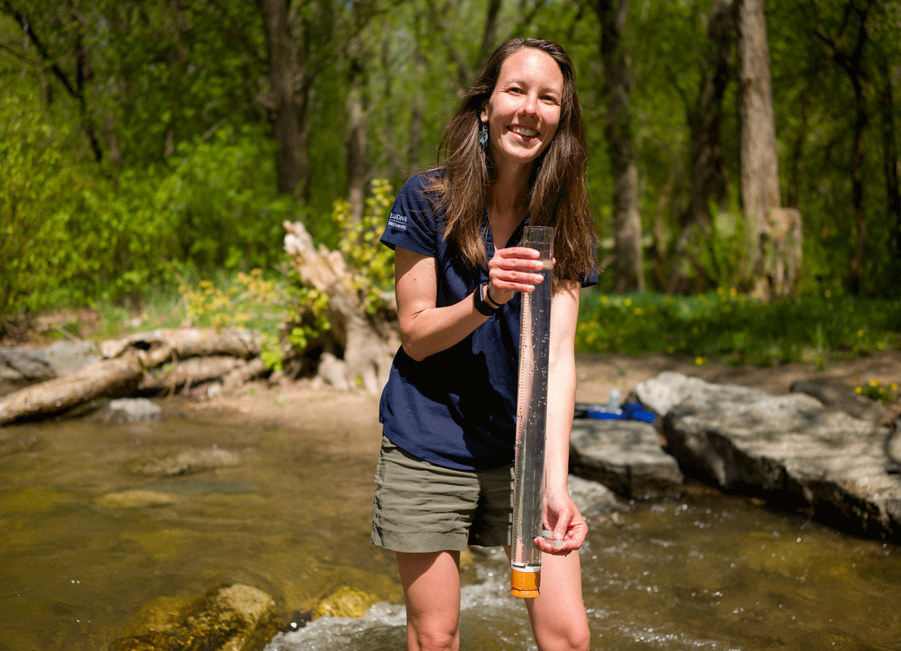 a woman stands in a stream holding a glass tube measuring device