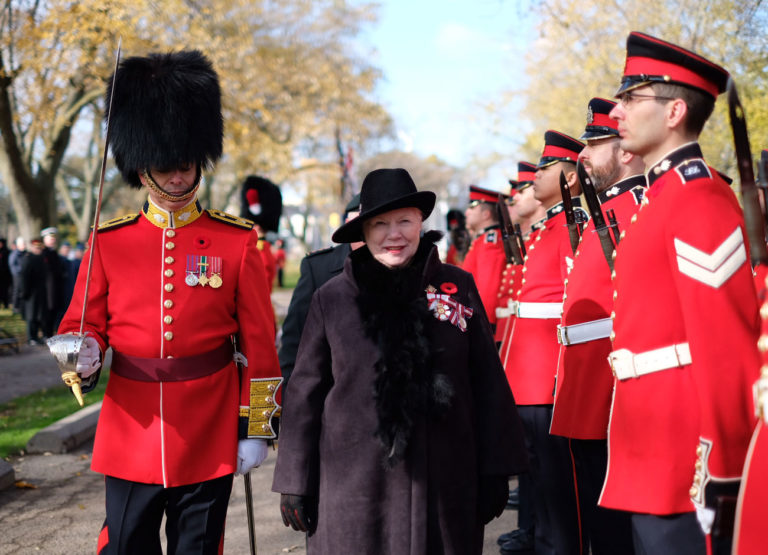 a woman dressed in a black coat and hat stands between guardsmen clad in red uniforms during a Canadian ceremony.