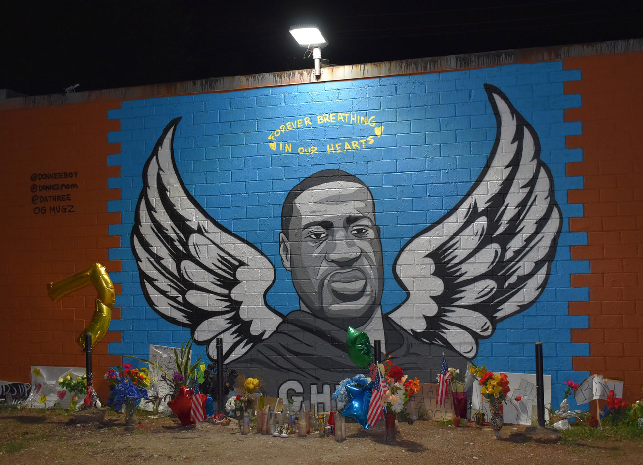 a memorial tribute to George Floyd. The mural depicts Floyd with angel wings and a halo painted on a brick wall. Flowers, stuffed animals. and signs from protests are on the ground. The mural is in Houston.