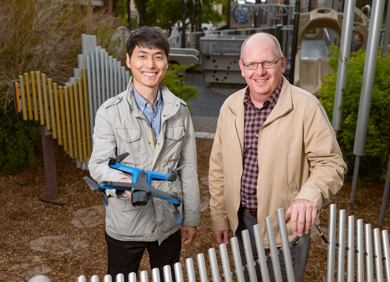 two professors stand in a playground, one holds a drone. Both are smiling. In the background are climbing structures, slides, and musical structures children activate through play.
