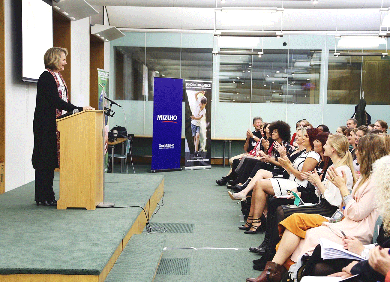 a blonde woman stands at a lectern while talking to an audience of women.