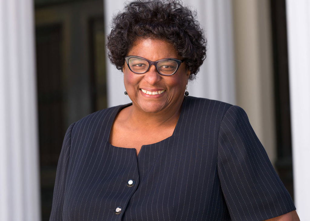 An African American woman in a navy pinstripe suit, dangly earrings, and sharp black glasses. smiles at the camera.