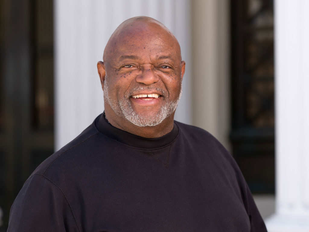 A bald African American man wearing a black shirt looks at the camera. He has white whiskers and is smiling.