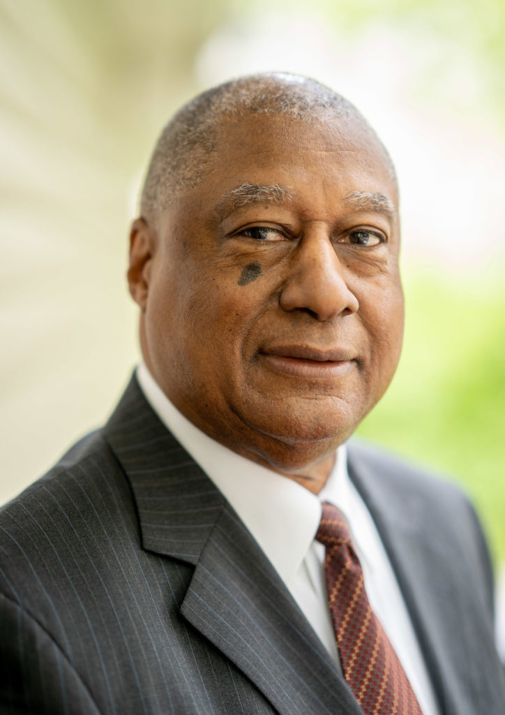 An African American man with salt and pepper hair and wearing a pinstripe suit and burgundy patterned tie half-smiles at the camera.