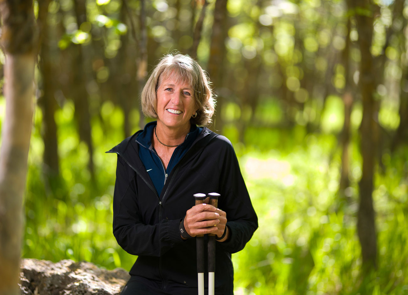 a woman in hiking gear holds a pair of hiking poles. She is sitting on a rock and smiling. She is surrounded by green trees with sunlight filtering through.