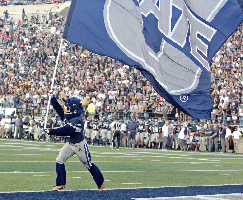 the Big Blue mascot runs across the football field waving a large blue Utah State flag behind him.