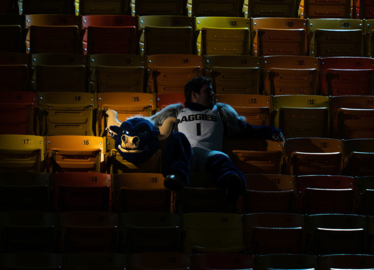a man sits in a mascot costume. He is holding the head on the seat next to him. His face is darkened by shadows. The stadium is empty.