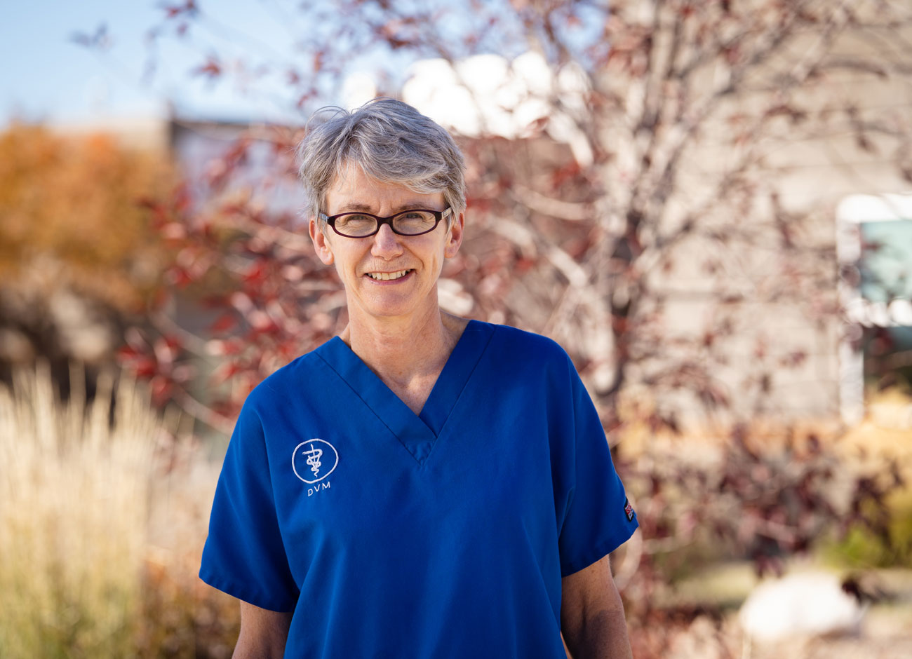 a woman with gray hair and black glasses smiles at the camera. She is wearing a royal blue veterinary technician outfit.