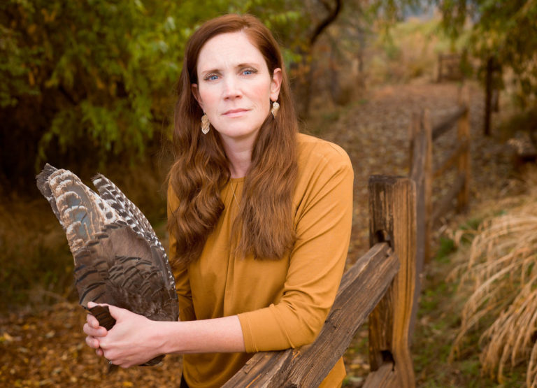 a woman with red hair and blue eyes holds a pair of hawk wings. She is leaning against a fence. She has a solemn look on her face.
