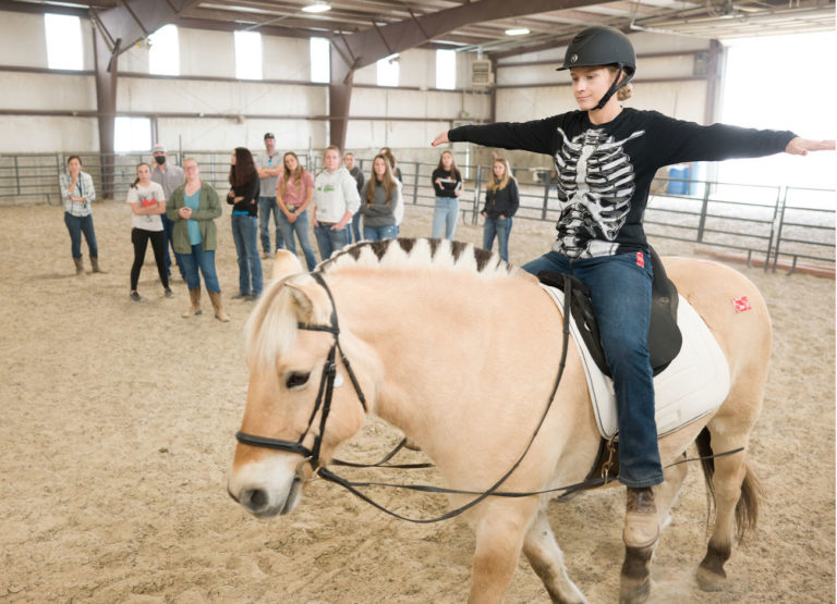 a woman in black shirt sits with her arms out on a horse during a therapy teaching session