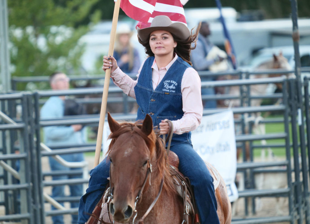 a young woman in a tan cowboy hat holds an American flag while riding her horse. She half smiles at the camera.