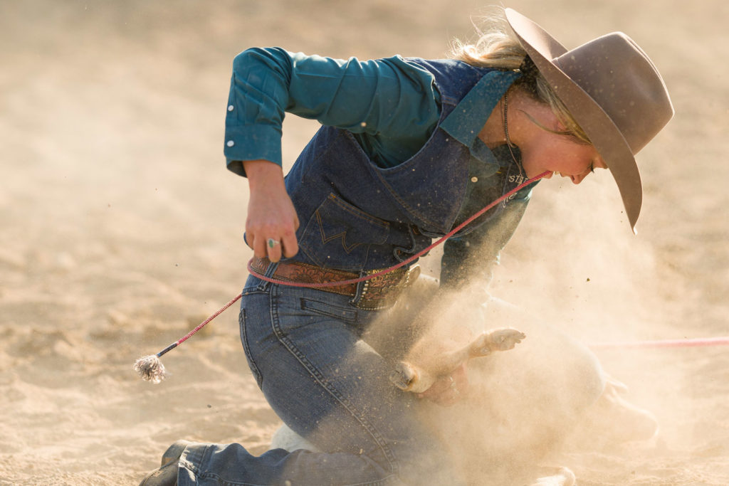 A young woman clad in denim ties a calf on the ground. A pink rope is between her teeth. Dust is flying, nearly making the calf invisible.