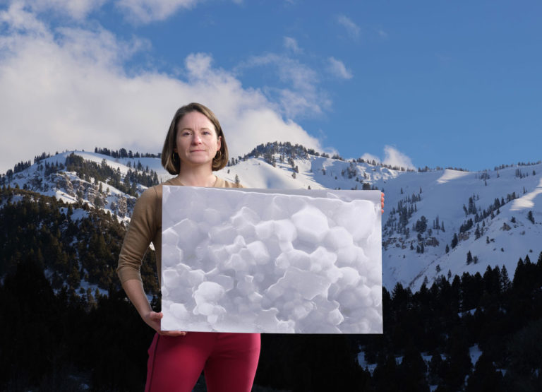 a woman in red pants holds a poster of hematite textures that look a little like clouds in front of a mountain range covered in snow.