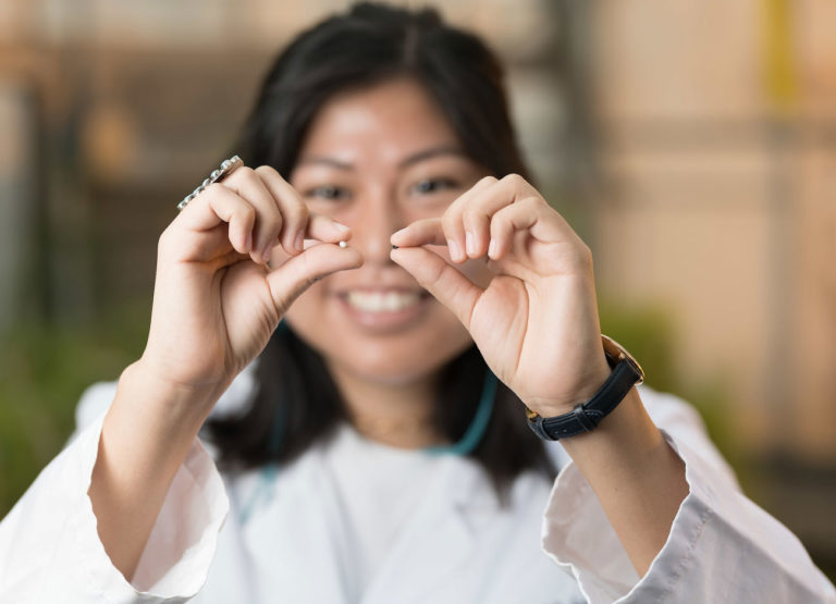 a young student holds two seeds between her hands - one white, one black. She is wearing a white lab coat and smiling.