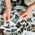 The tips of white boots stand near a pile of polaroids showing the feet and pants and shorts of dozens of people. Above them are two hands hold the edges of a polaroid of a woman's skinny jeans and white sandals.
