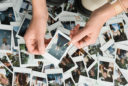 The tips of white boots stand near a pile of polaroids showing the feet and pants and shorts of dozens of people. Above them are two hands hold the edges of a polaroid of a woman's skinny jeans and white sandals.