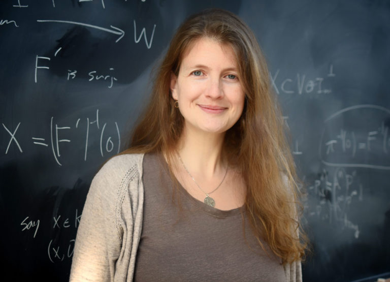 a woman with long blonde hair and tan shirt and sweater stands in front of a black board with physics equations written in chalk. She has a knowing smile.