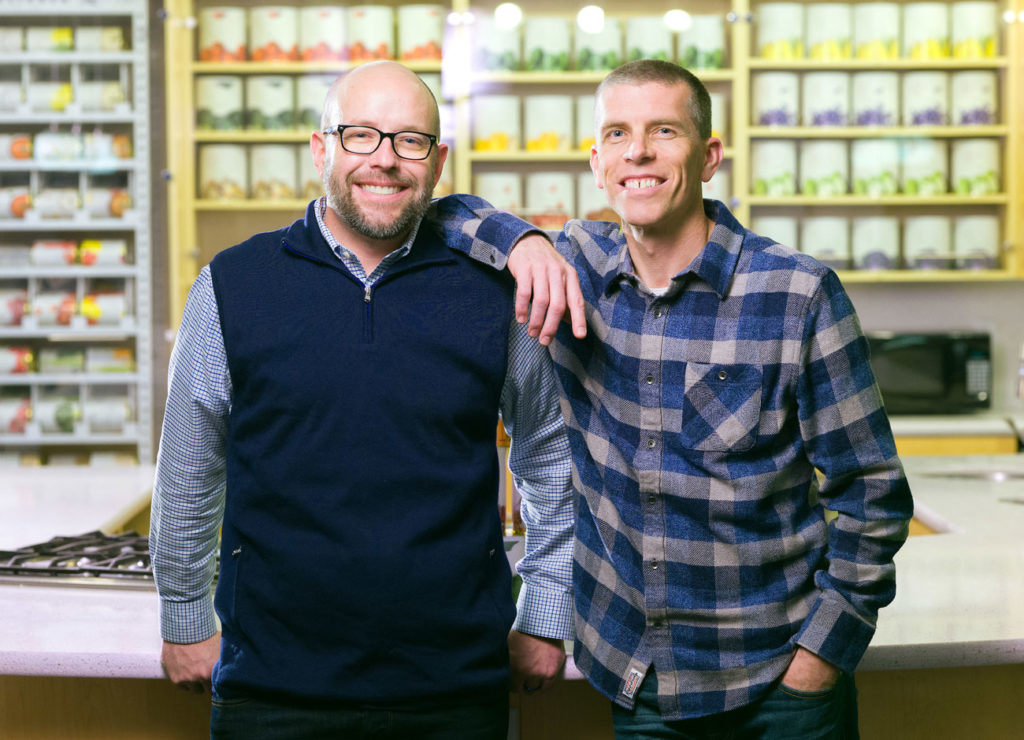 Two men lean toward one another shoulder to shoulder while standing in front of a display case of canned freeze dried vegetables and fruits.