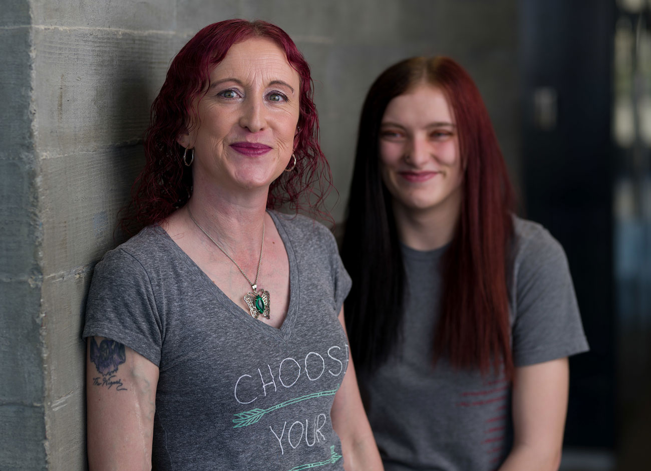 a woman with red hair and blue eyes smiles with her mouth closed. She has a butterfly necklace and shirt that reads "Choose Your" with arrows pointing in different directions