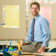 a man in a blue button up shirt and tie sits on his desk in a school classroom, surrounded by small robots he teaches students to build.