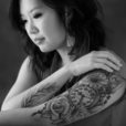 A young Asian woman gracefully displays her tattoos, one which has a clock referencing the time of her birth on one arm. The other arm has a Buddhist symbol of compassion