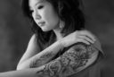A young Asian woman gracefully displays her tattoos, one which has a clock referencing the time of her birth on one arm. The other arm has a Buddhist symbol of compassion