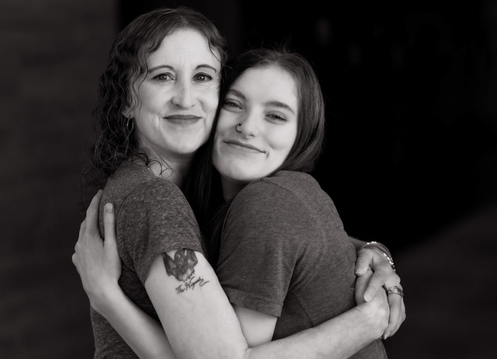 A woman and her daughter hug with their cheeks pressed against each other. They smile in the camera.