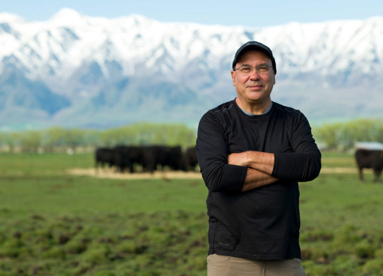 Juan Vilalba stands in a bright green field with snow capped peaks in the background. Cattle graze behind him.