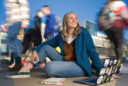 a young woman sits in the middle of a busy campus. People whiz behind her in multiple blurs. She has her laptop open and is looking off with a confident look on her face.