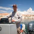 Dan Keller stands at the helm of a boat, puffy white clouds and a blue sky behind him. The water of Lake Powell gleams in the background.