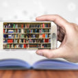 a hand holds a smartphone showing a library of books on the screen. In the background is a textbook open on a table.