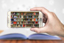 a hand holds a smartphone showing a library of books on the screen. In the background is a textbook open on a table.