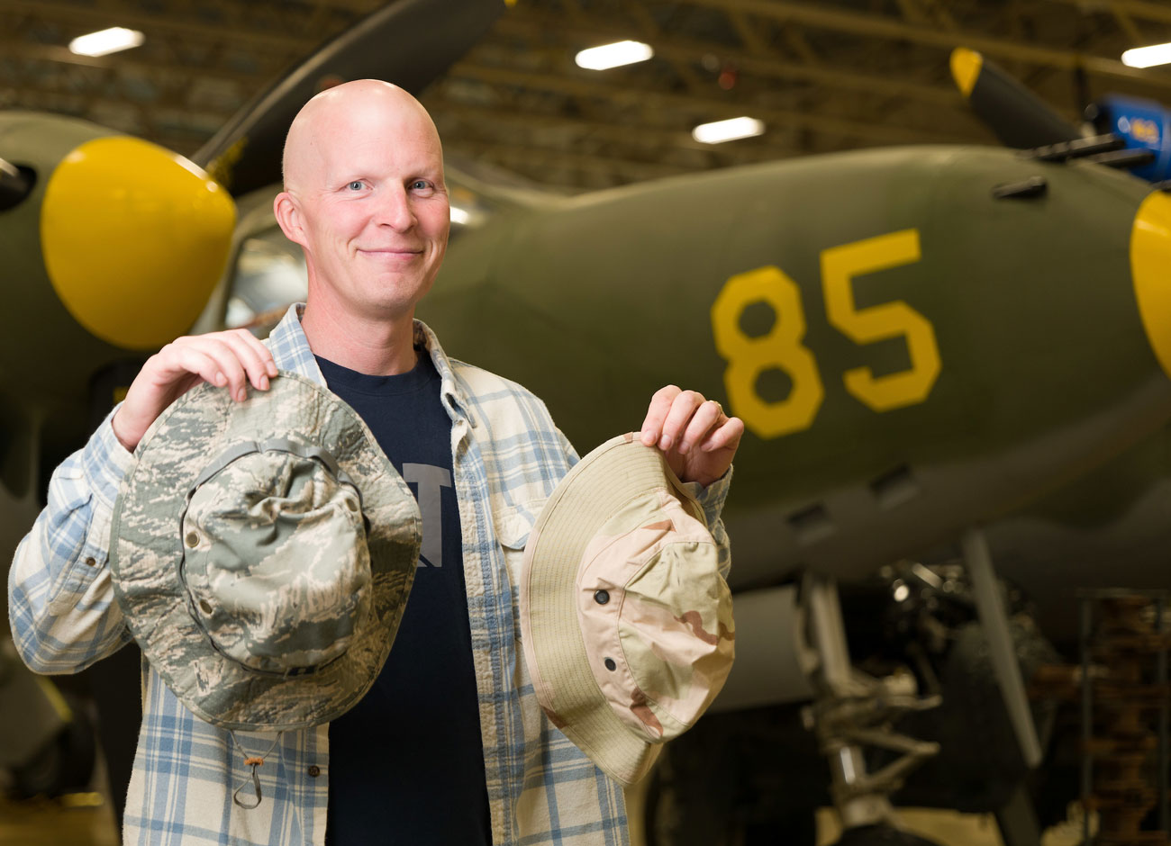 a man holds two camouflage hats from the War in Iraq. Behind him is a green airplane with the number 85 painted on the side.