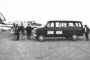 a black and white image of a van with Utah State University written on the side with a small cluster of professors heading to a small aircraft in the background