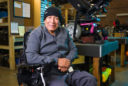 a man in a gray button up sweater and black beanie sits in a wheelchair inside USU's assistive technology workshop
