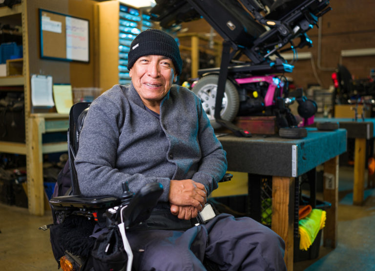 a man in a gray button up sweater and black beanie sits in a wheelchair inside USU's assistive technology workshop